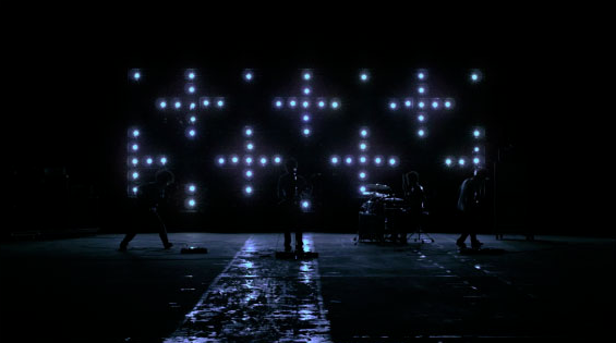 Band on sound stage playing in the dark with lights in background shaped like plus signs
