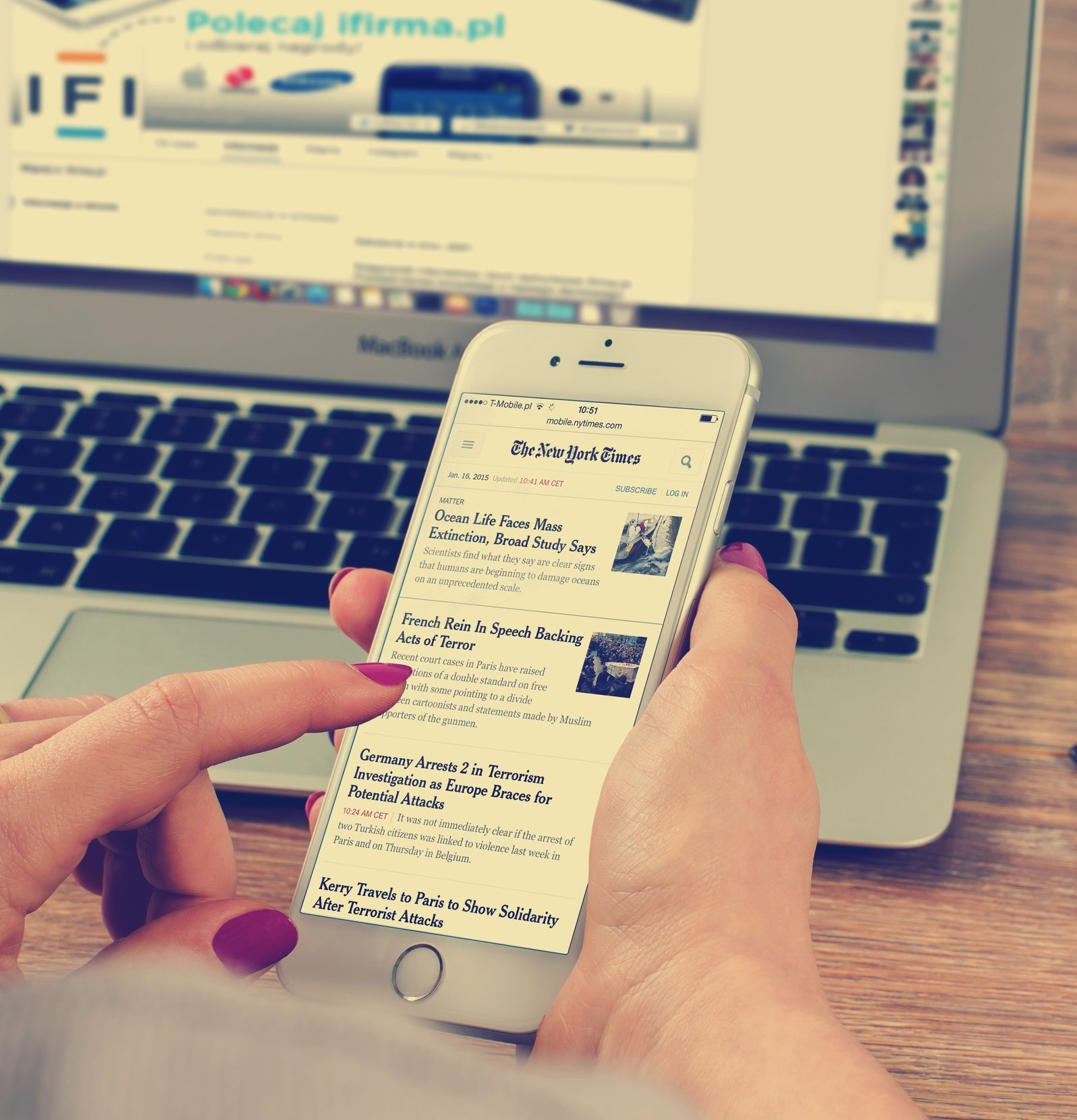 Image of a female hands holding an iPhone, reading The New York Times and a MacBook laptop