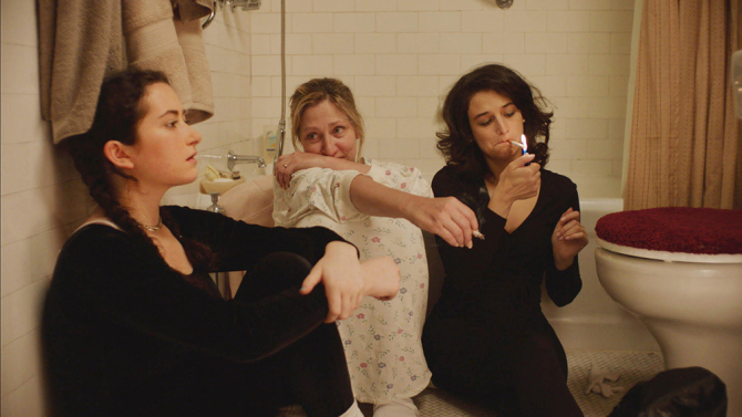 Film still from Landline. Edie Falco sits on the floor of a bathroom smoking with Jenny Slate and Abby Quinn.