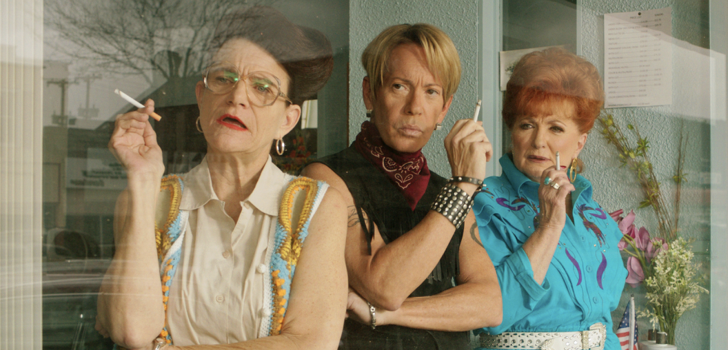 Two older women and one middle aged male hairdresser peering through a window and smoking