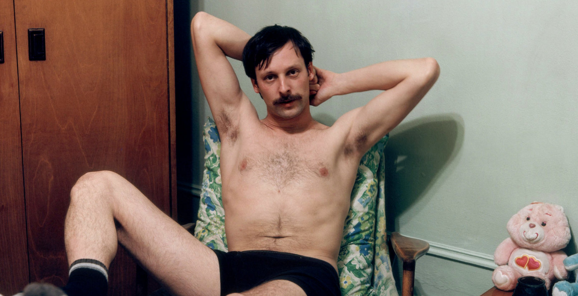 Mustached man in black underwear and socks lounging on a chair. A plush Care Bear is next to him.