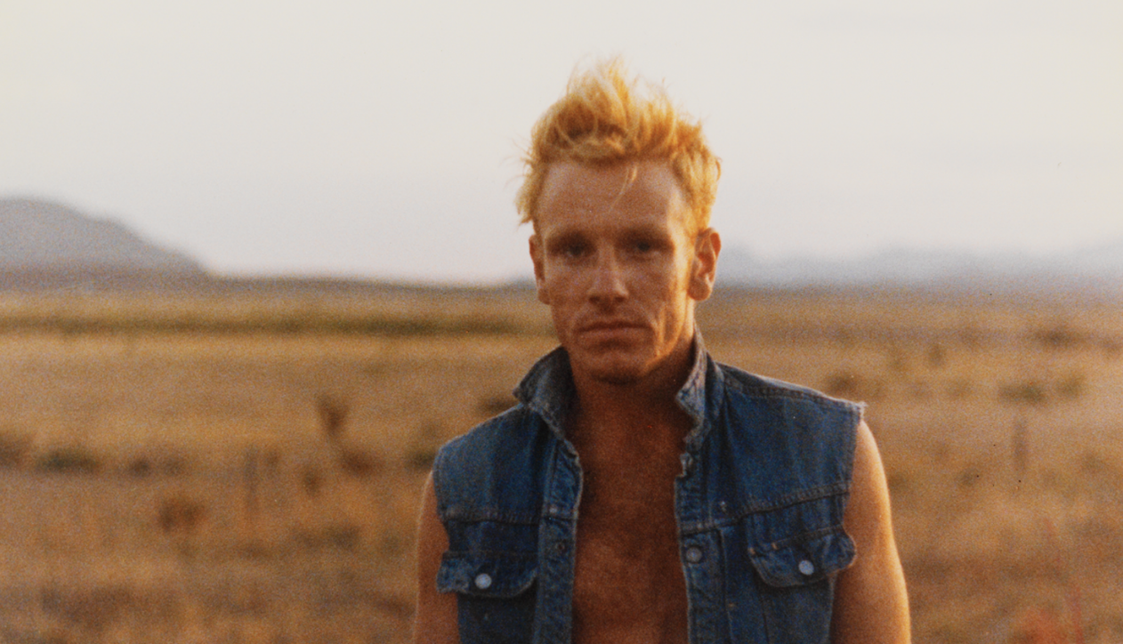 Young man shot from chest up. He's only wearing an open denim vest. His hair is messy and red. He stands on an open plain.