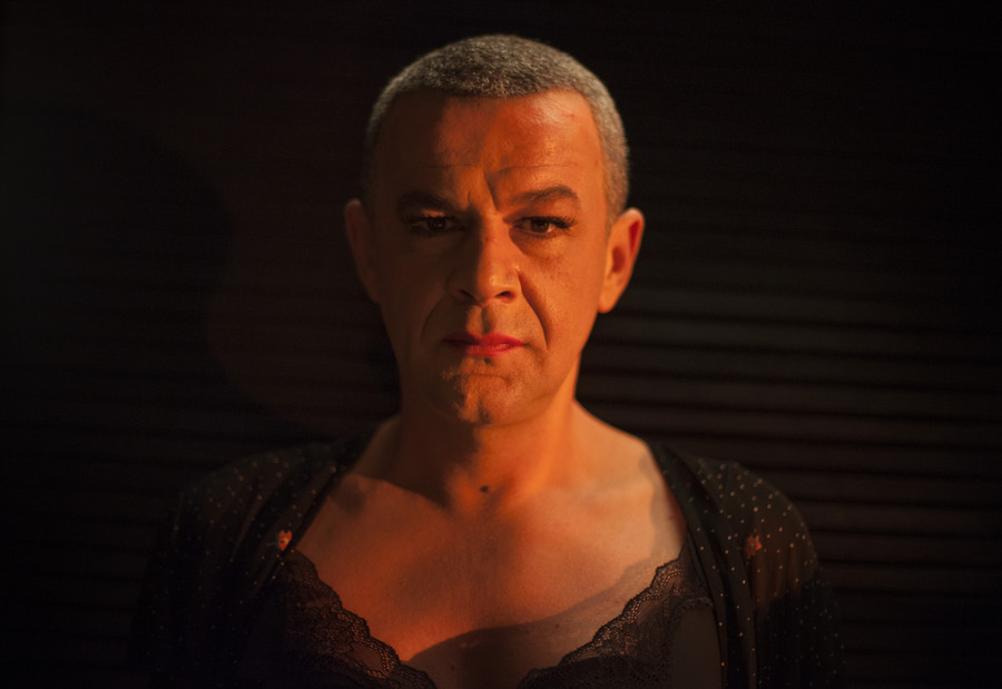 A middle aged white man is seen from chest up. He has short hair and wears makeup and lingerie
