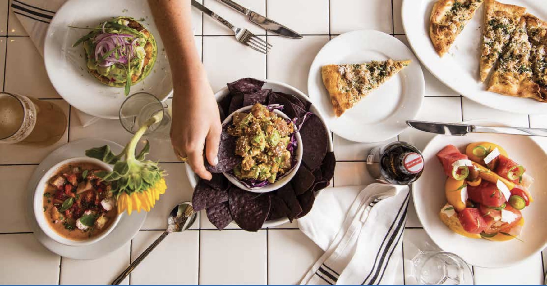 Food on a table, with a person's hand dipping a chip into guacamole