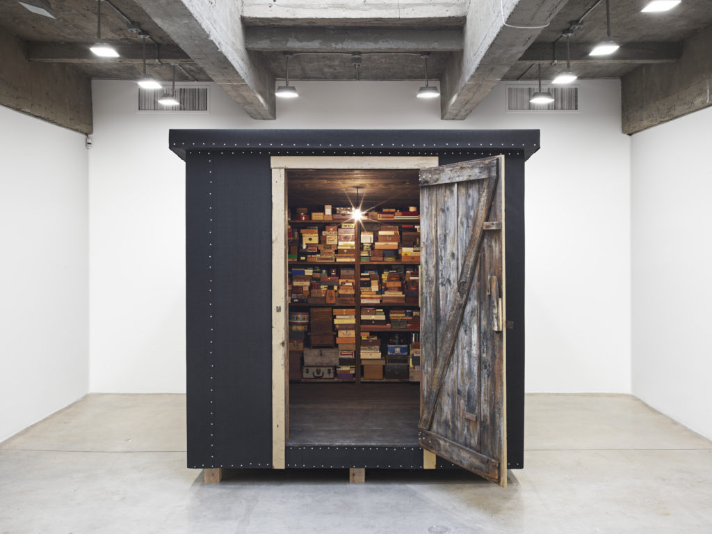 An art exhibition in a white-walled gallery featuring a black shed, door ajar, showing a bunch of neatly stacked boxes on shelves inside the shed