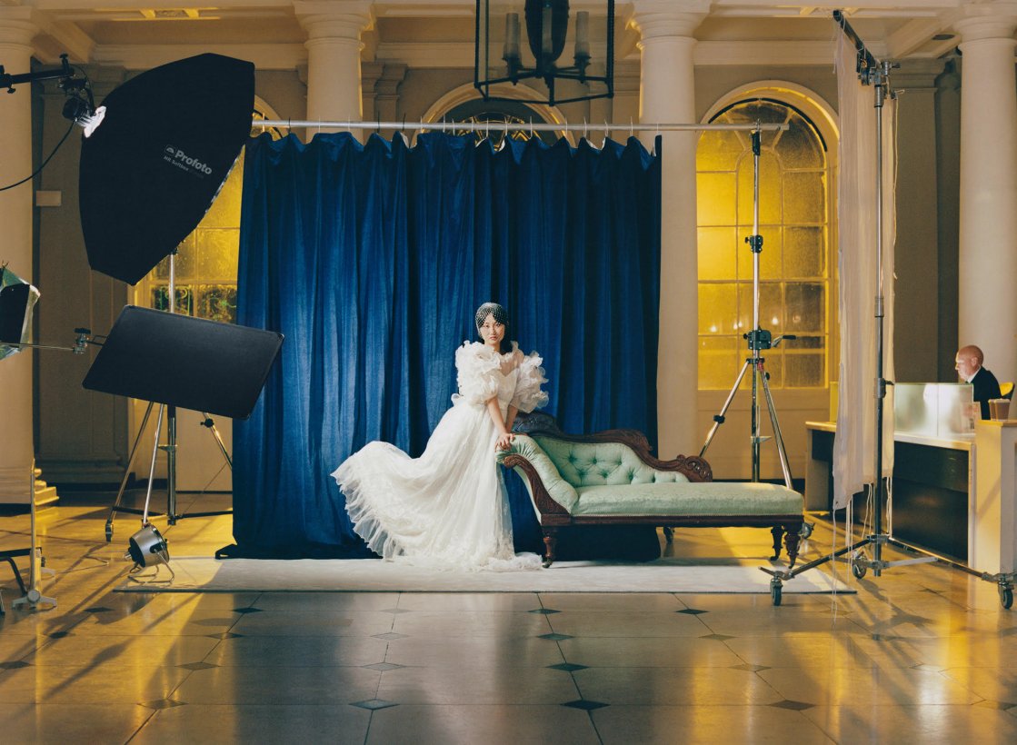 A young Asian woman in a white wedding dress poses in a hotel lobby leaning against an antique green fainting couch. She looks to be in a photoshoot, with photography equipment around her, and she's in front of a blue drape
