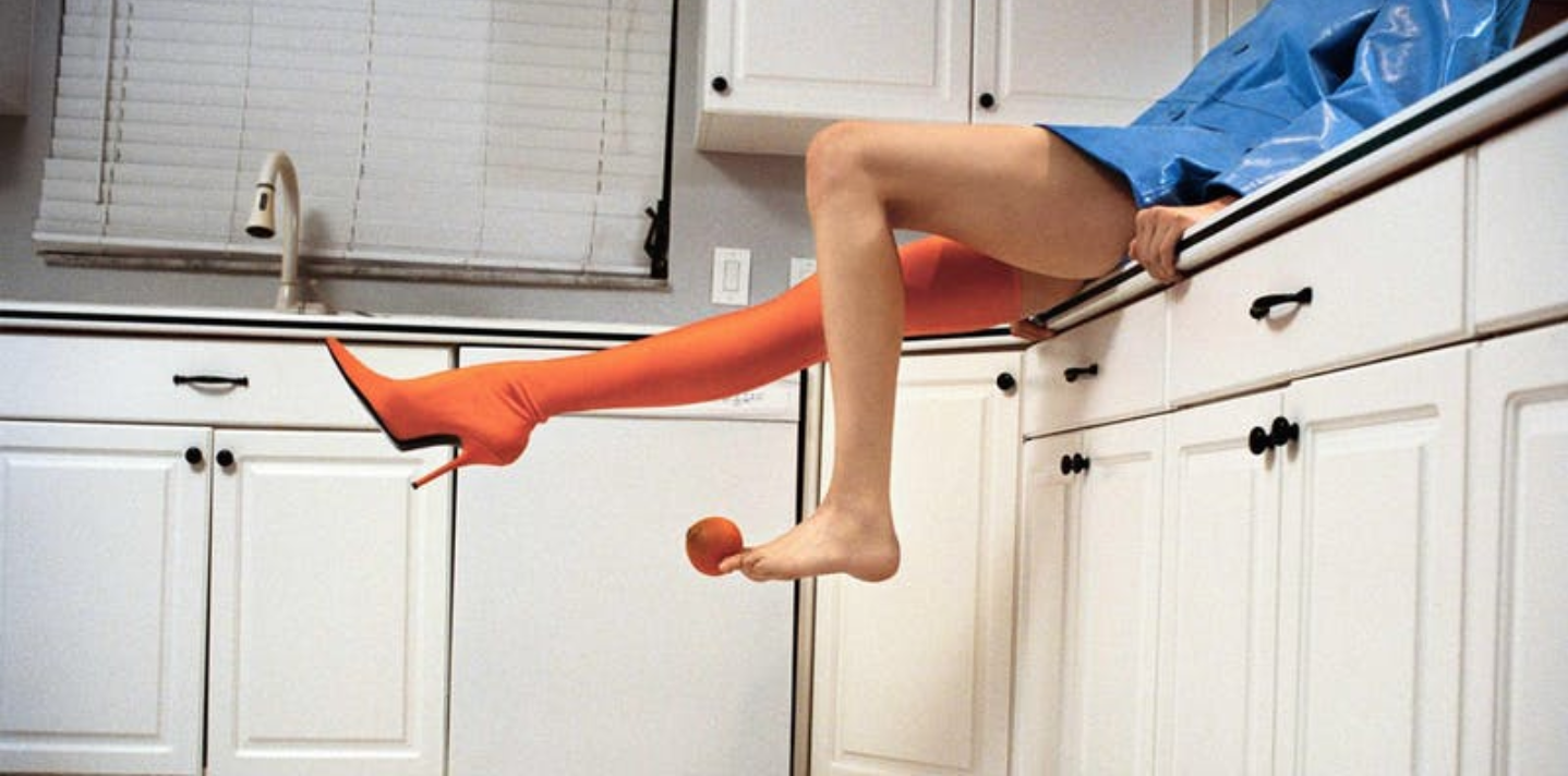 Image of a someone sitting on a kitchen counter. You can see the person is wearing a blue raincoat, no pants, and the right leg has a knee-high orange boot with a spike heel