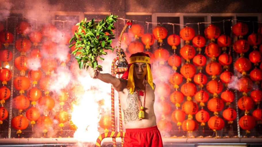 Shirtless, tattooed Asian man with leaved branches in his extended right arm, standing in front of a wall of red Chinese lanterns