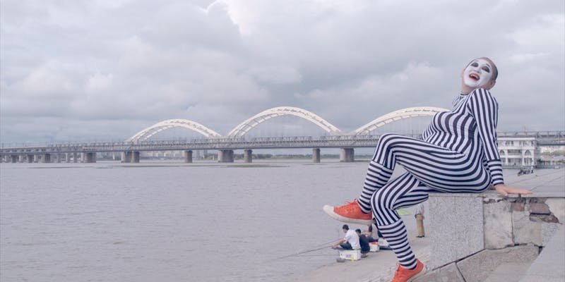 A person dressed as a mime sits beside a waterway, with a bridge in the background