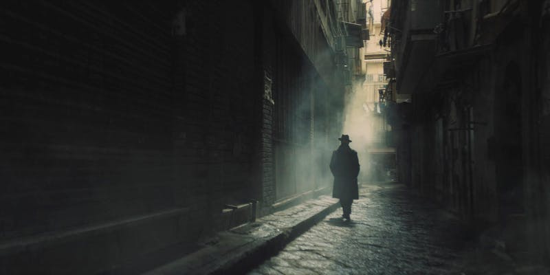 Man in hat and trenchcoat walking down a dark and shadowy alley