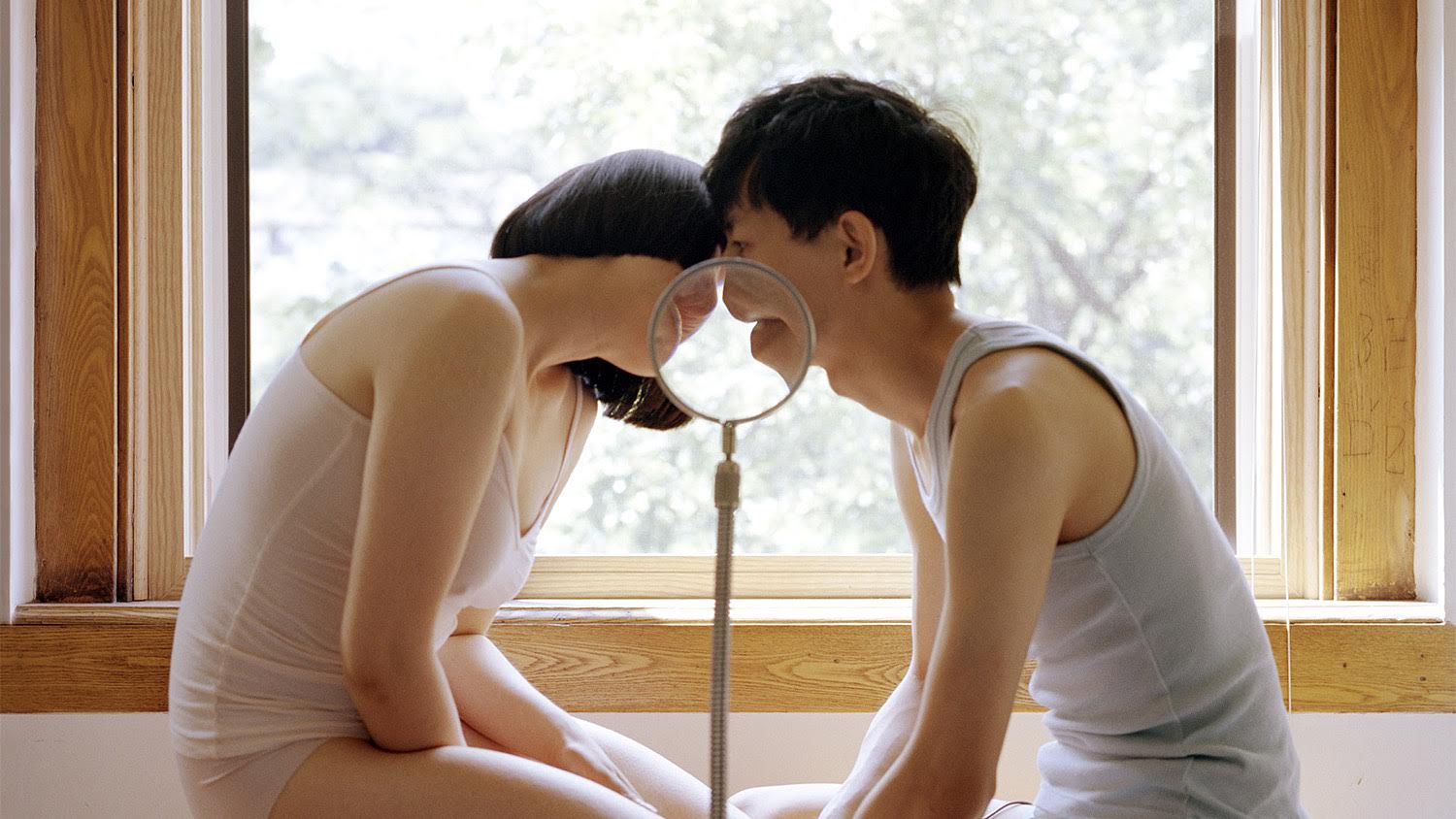 Image of an Asian woman and Asian man in white underwear seated on the floor and kissing, with a magnifying glass magnifying their lips