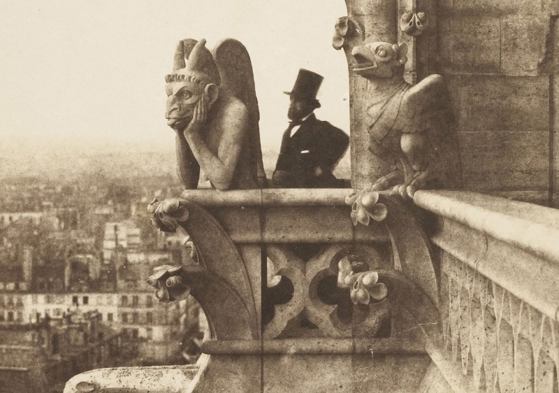 An old sepia image of a bearded man in a top hat standing on a parapet of a tall building beside a large gargoyle