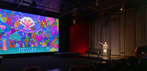 A person points to an animated image shown on screen in a theater.